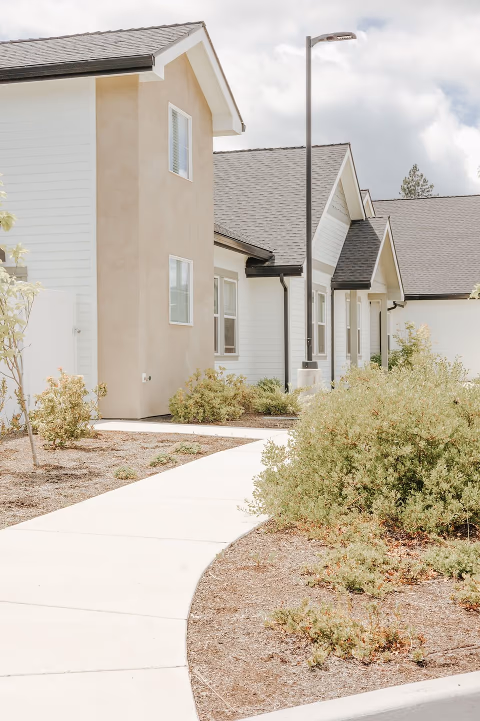 Exterior view of a senior living facility building with beige and white walls, multiple windows, a sloped roof, a streetlamp, and a curved concrete walkway surrounded by landscaping with bushes and small trees.