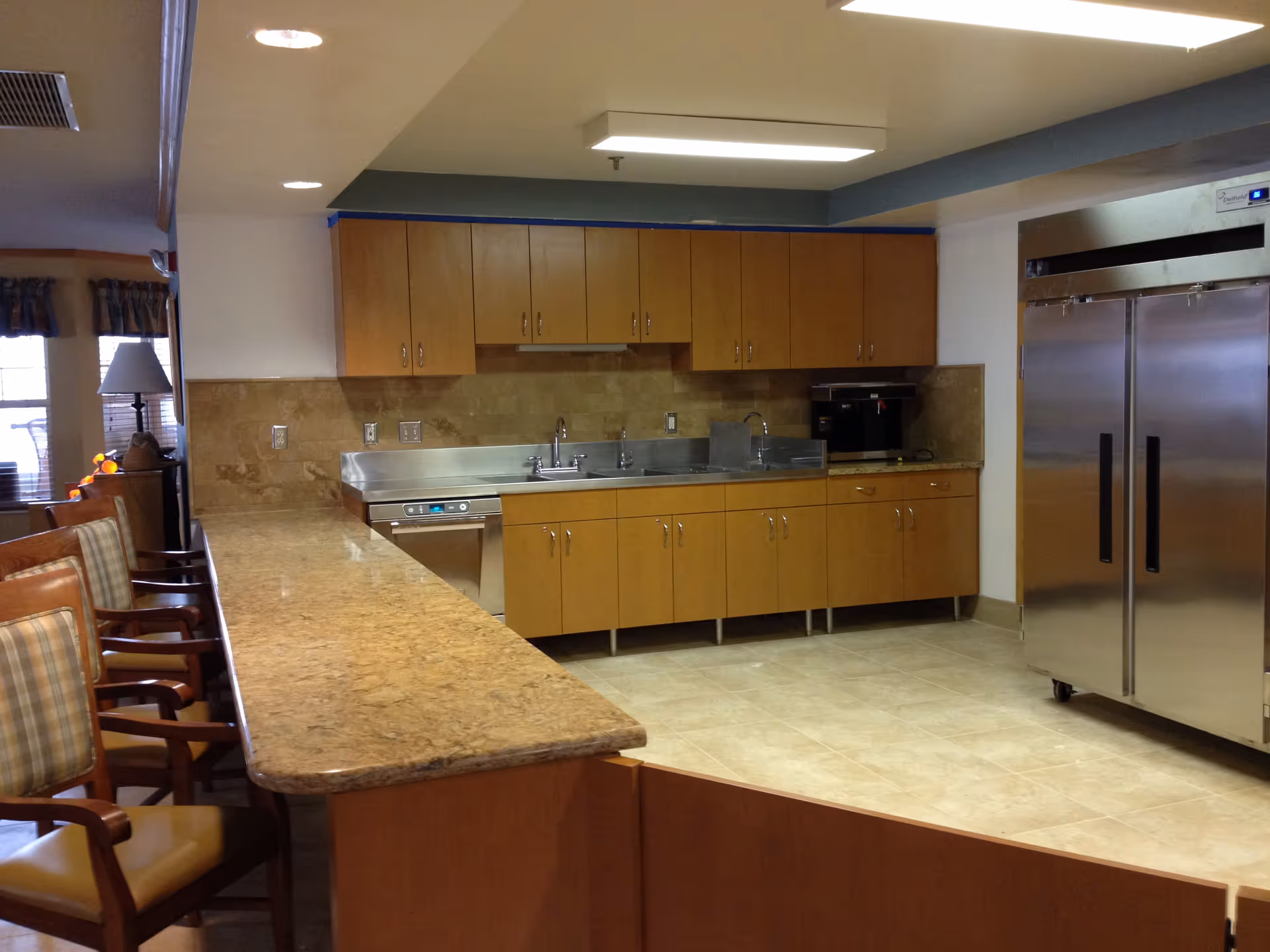 Interior view of a kitchen area in a senior living facility with wooden cabinets, a stainless steel double-door refrigerator, a coffee machine, a dishwasher, and a long granite countertop with several chairs lined up along it. The kitchen has tiled flooring and recessed lighting.
