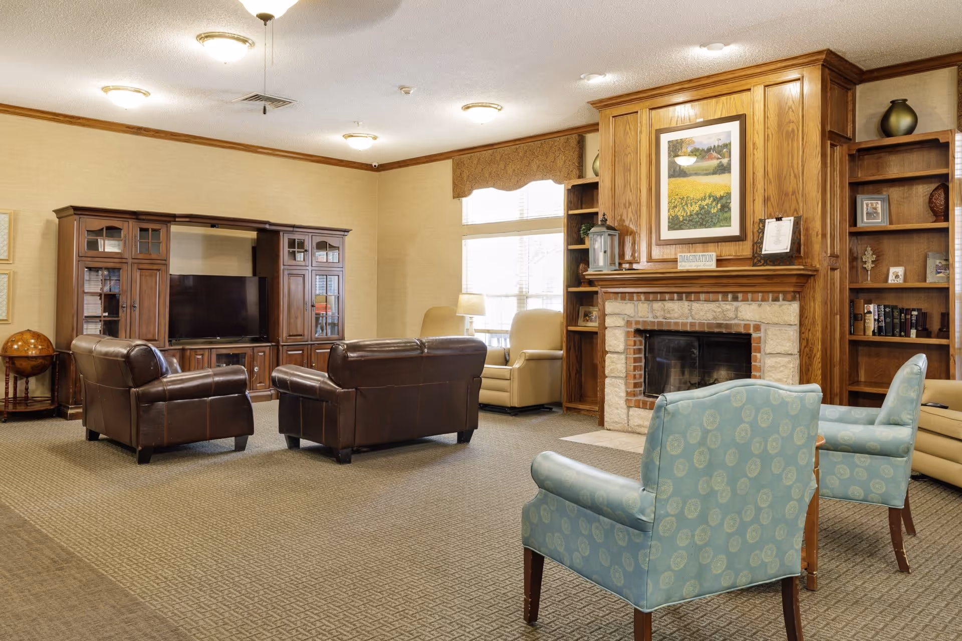 A cozy living room with a stone fireplace surrounded by wooden built-in shelves filled with books and decorative items. There are two blue patterned armchairs in the foreground, two brown leather armchairs and a matching loveseat facing a large wooden entertainment center with a flat-screen TV. The room has beige walls, carpeted floors, and large windows letting in natural light.