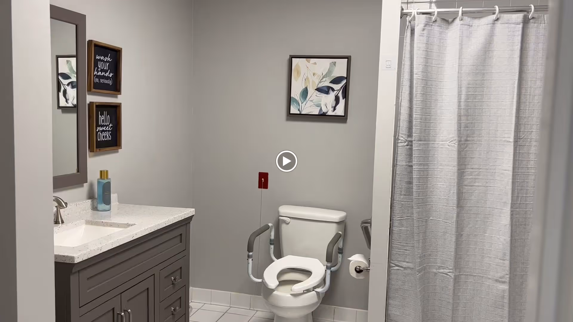 A clean bathroom with a white toilet equipped with safety rails, a white shower curtain, a gray vanity with a white countertop and a sink, a mirror above the sink, and two framed signs on the wall that read 'wash your hands (no, seriously)' and 'hello sweet cheeks'.