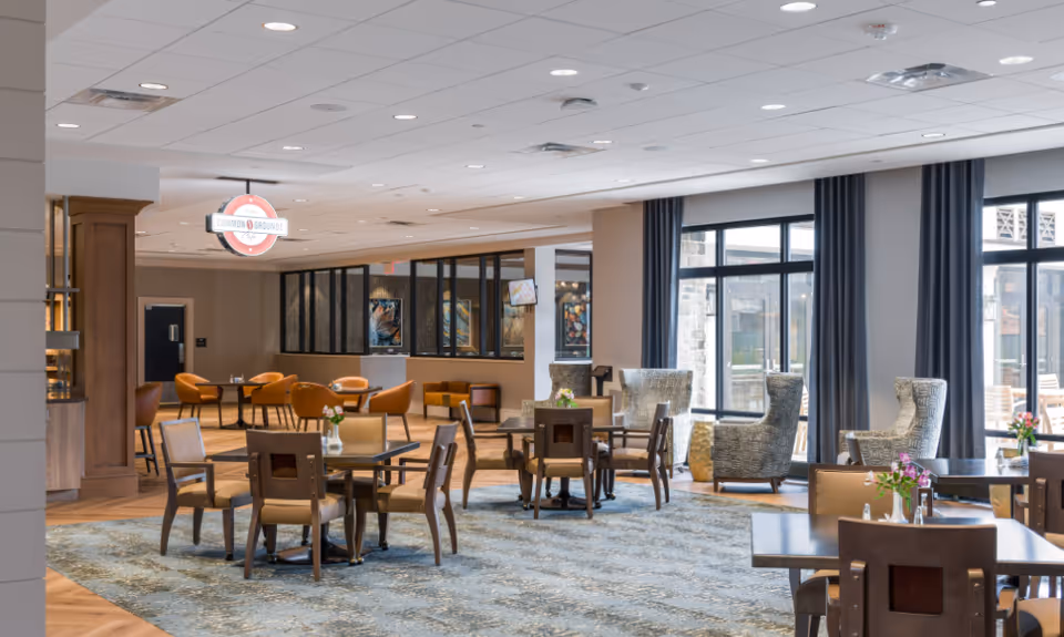A spacious and well-lit dining area in a senior living facility with multiple tables and chairs arranged neatly. The room features large windows with dark curtains allowing natural light to enter. There are comfortable armchairs near the windows and a sign hanging from the ceiling that reads 'Common Grounds Cafe'. The flooring is a combination of wood and carpet, and the overall decor is modern and inviting.