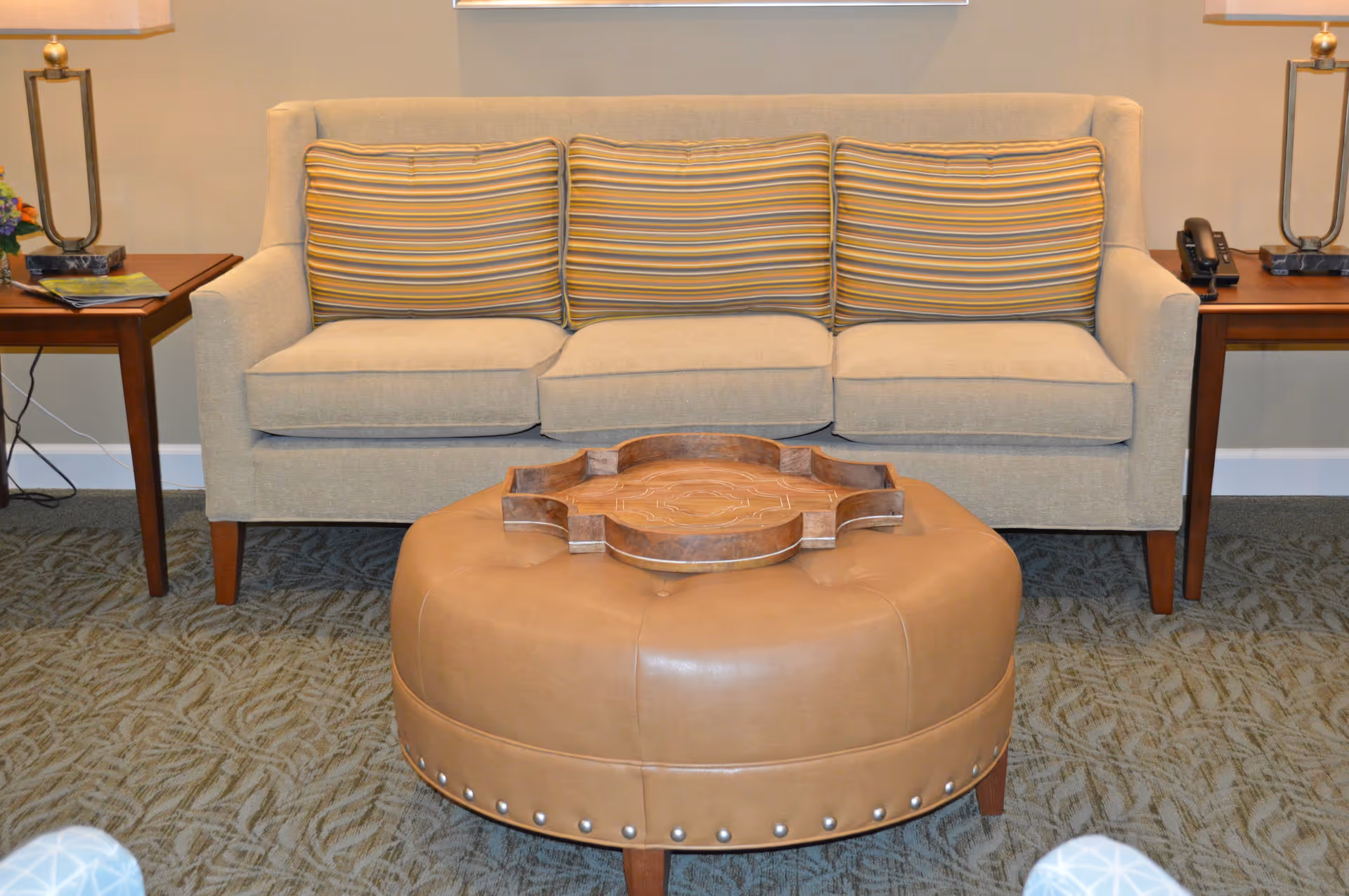 Beige three-seat sofa with striped cushions flanked by wooden side tables and a round leather ottoman with a decorative tray in front.