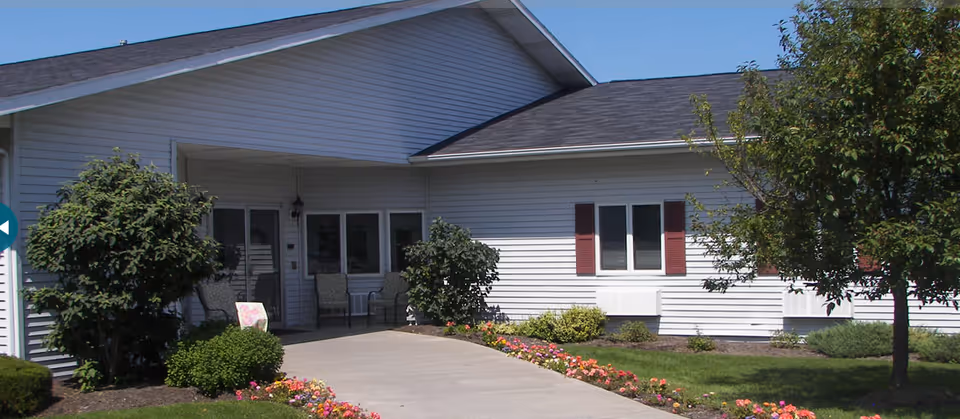 Exterior view of a single-story building with white siding and a dark roof, featuring a covered entrance with chairs and a walkway bordered by colorful flowers and green shrubs.