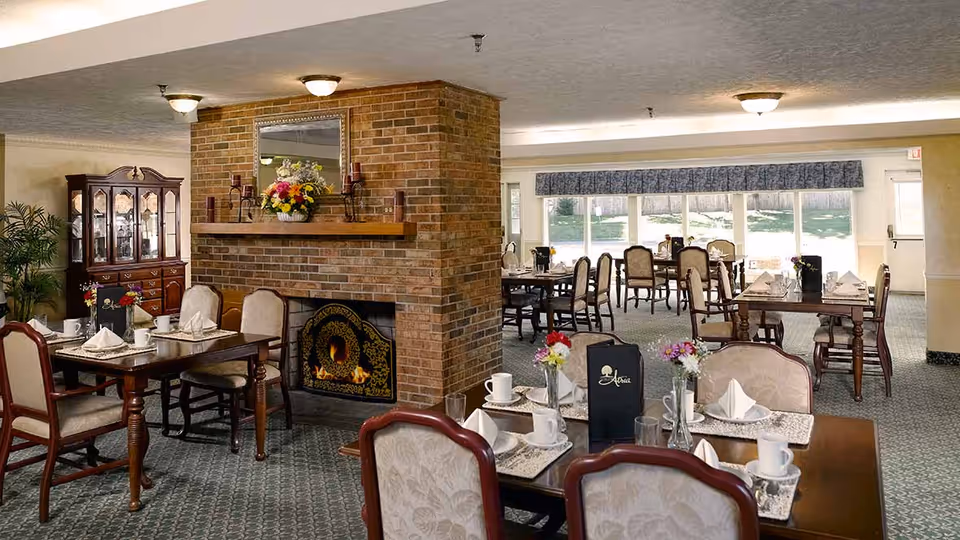 A senior living community dining room with multiple wooden tables and upholstered chairs arranged neatly. Each table is set with white napkins, cups, glasses, and small floral centerpieces. A brick fireplace with a decorative screen and a mirror above it is centrally located, adding a cozy ambiance. Large windows in the background allow natural light to fill the room.