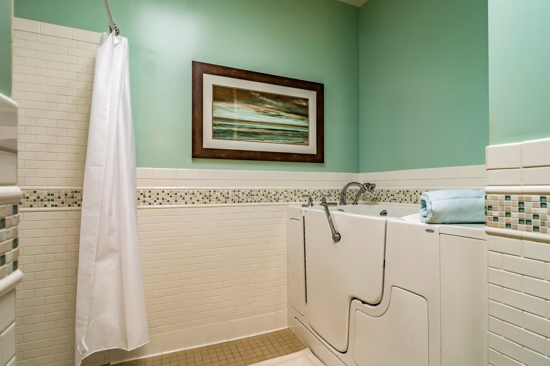 A bathroom with light green walls and white tiled lower walls featuring a mosaic border. There is a walk-in bathtub with a door and a folded blue towel on its edge. A white shower curtain hangs on the left side, and a framed picture of a seascape is mounted on the wall above the bathtub.