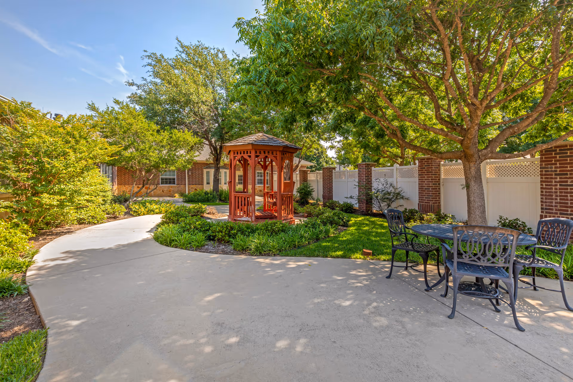 A peaceful outdoor garden area with a curved concrete pathway, a wooden gazebo surrounded by greenery, and a black metal table with four chairs under a large tree. The area is enclosed by a white fence with brick pillars and there is a brick building in the background.