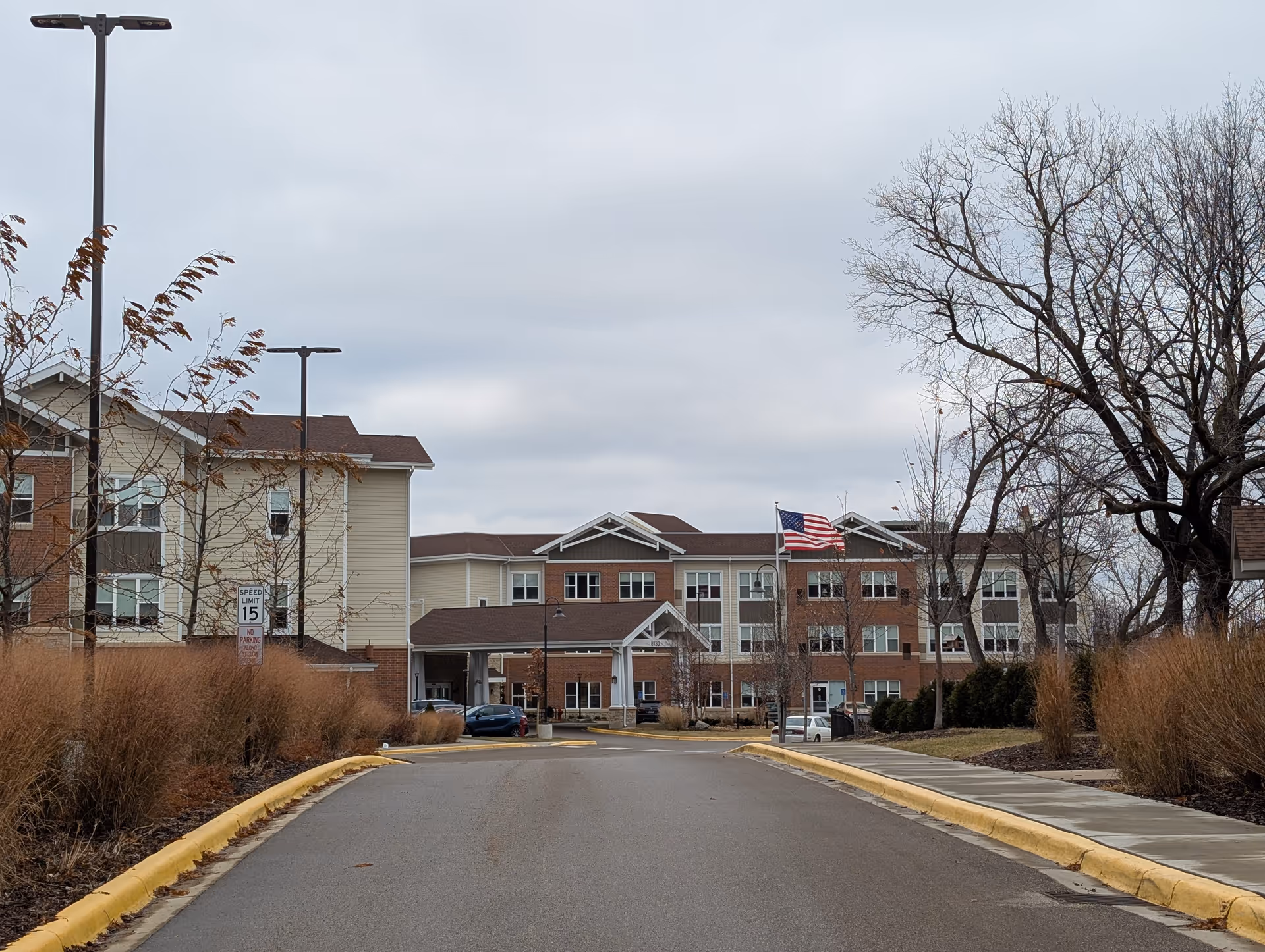 View of the front exterior of a senior living facility building with a driveway leading up to the entrance. The building has three stories with a combination of brick and beige siding. There are leafless trees and brown ornamental grasses on either side of the driveway. An American flag is flying on a flagpole near the entrance under a cloudy sky.