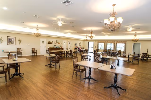 A spacious dining room with wooden floors and multiple wooden tables and chairs arranged throughout. The room is well-lit with ceiling lights and chandeliers, and there are large windows along one wall letting in natural light. The walls are decorated with framed pictures and sconces.