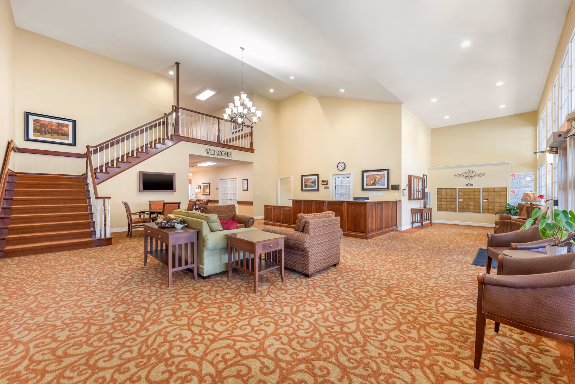 Spacious senior living facility lobby with patterned carpet, multiple seating areas including sofas and armchairs, a wooden reception desk, staircase with wooden railing, and large windows letting in natural light.