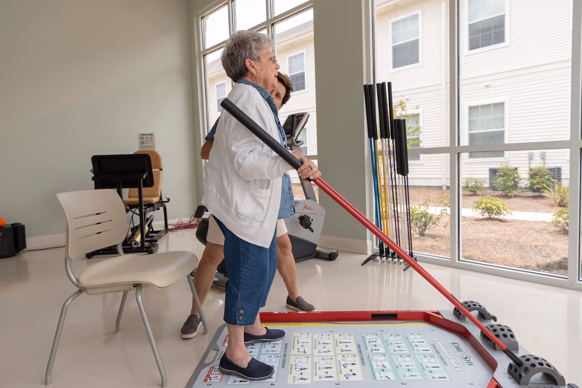 An elderly woman is standing on a balance board holding a long stick, assisted by a caregiver in a bright room with large windows. Exercise equipment and a chair are visible in the background.