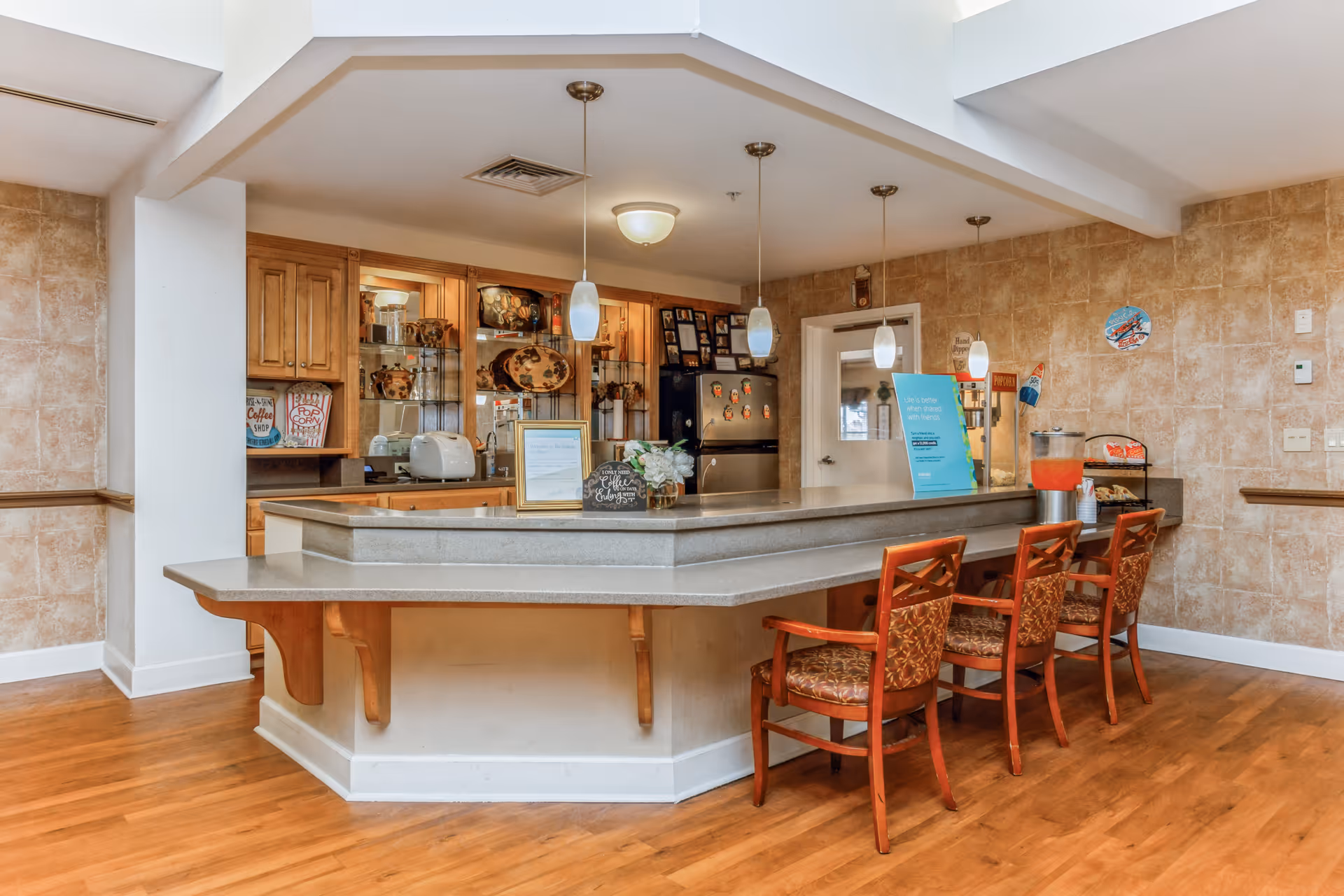 A cozy kitchen area in a senior living facility with a large counter and three wooden chairs with patterned cushions. The counter has a flower vase, framed sign, and a blue informational sign. Behind the counter, there are wooden cabinets, a refrigerator with magnets, a toaster, and a popcorn machine. The walls are tiled with a beige pattern, and the floor is wooden.