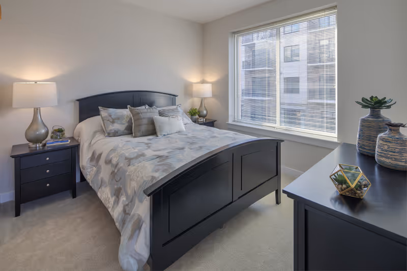 A well-lit bedroom with a black wooden bed frame and a bed made with floral-patterned bedding and multiple pillows. There are two matching black nightstands on either side of the bed, each with a modern lamp. A large window with blinds lets in natural light, and a black dresser with decorative vases and a small geometric terrarium is positioned near the window.