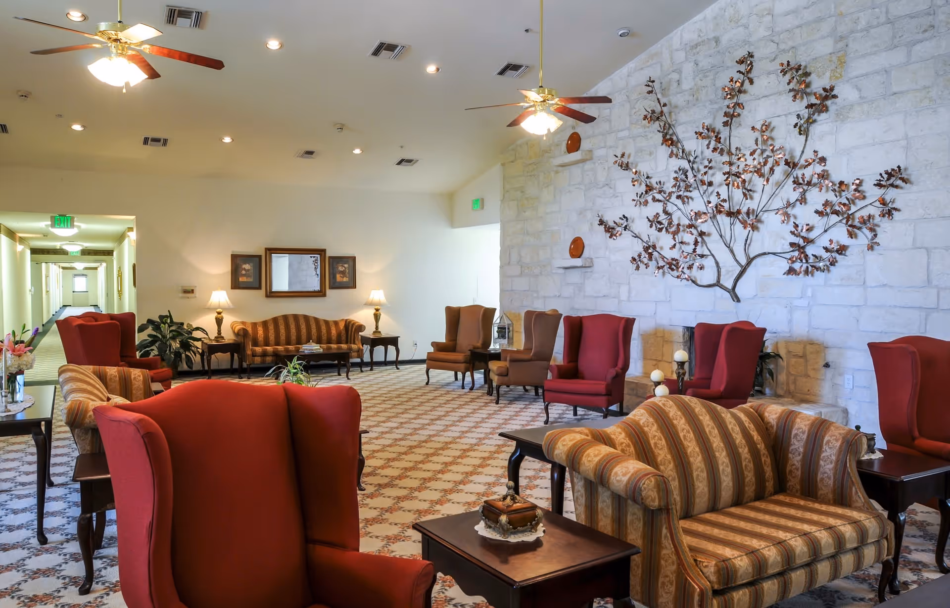 Spacious senior living lounge with upholstered armchairs and sofas arranged around side tables and a stone accent wall decorated with metal branches.