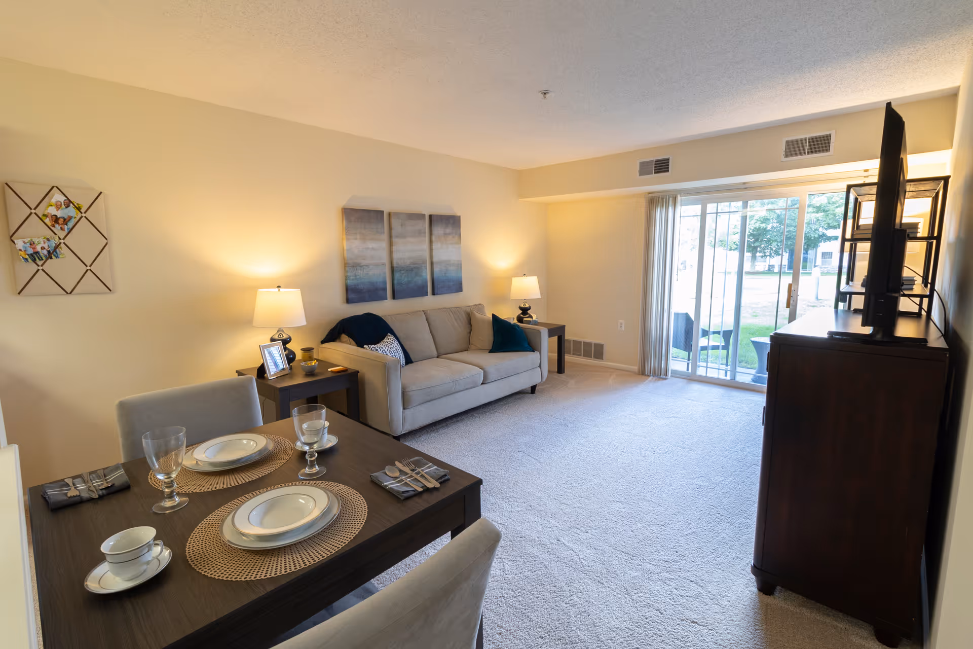 A bright living room and dining area in a senior living facility apartment. The living room features a beige sofa with cushions, two side tables with lamps, and a TV on a dark wooden stand. The dining area has a dark wooden table set for two with plates, glasses, and napkins. Large sliding glass doors lead to an outdoor patio with chairs and a table, allowing natural light to fill the room. The walls are light-colored, and there is carpet flooring throughout.