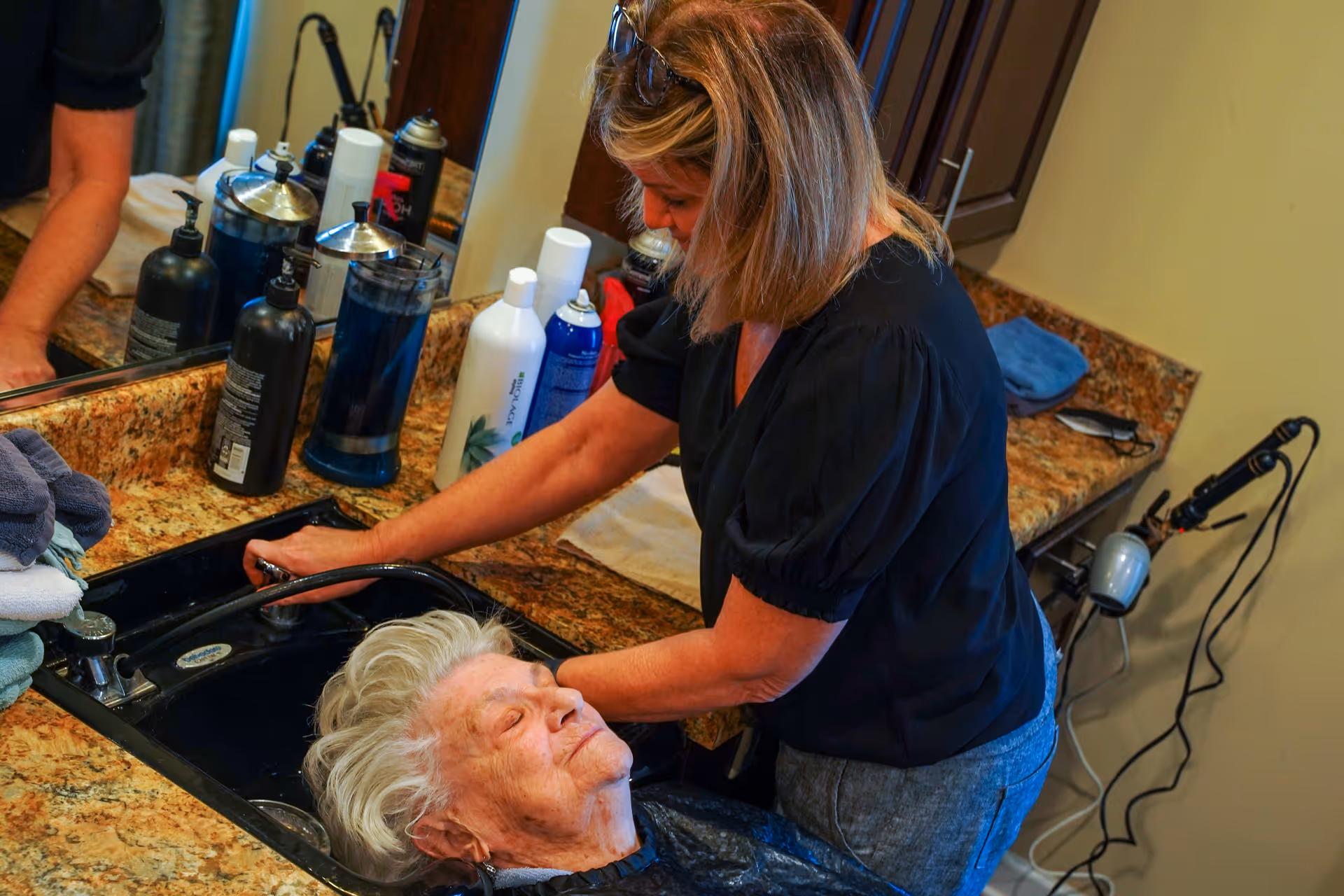 A caregiver washes an older woman's hair over a sink at a salon-style countertop with bottles and a mirror.