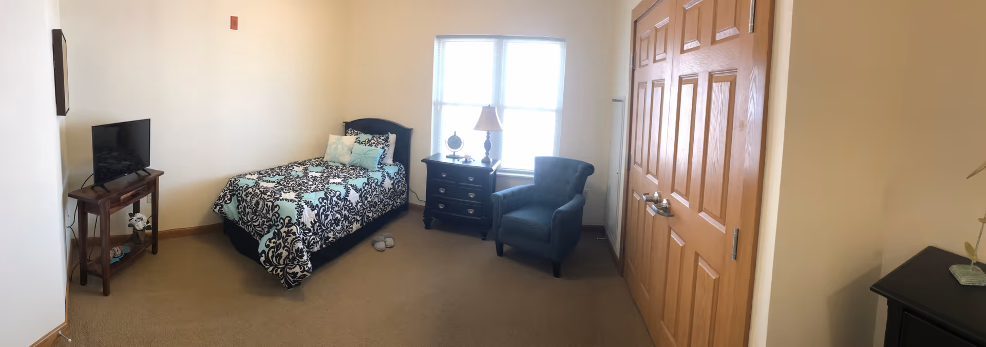 A small bedroom with a single bed covered in a black and white patterned comforter with light blue accents. Next to the bed is a dark wooden nightstand with a lamp and decorative items. A blue upholstered armchair is placed near a window with white blinds. A wooden TV stand with a flat-screen TV is positioned against the opposite wall. The room has beige carpet and light-colored walls, with wooden closet doors on one side.