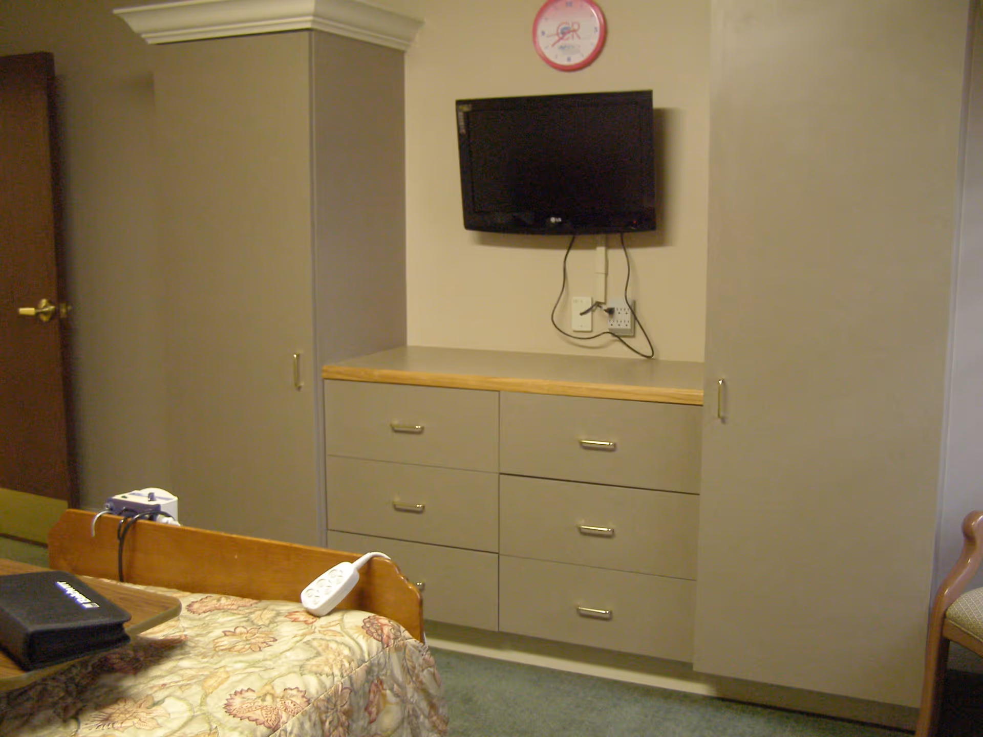 Interior view of a senior living facility bedroom featuring a bed with a floral bedspread, a mounted flat-screen TV above a set of six drawers with a wooden countertop, and two tall cabinets on either side. A clock is mounted on the wall above the TV, and a chair is partially visible on the right side.