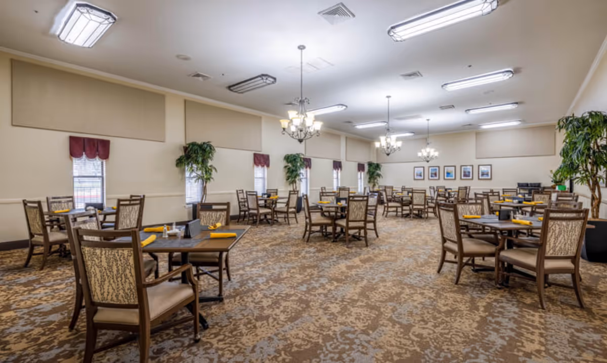 A spacious dining room with multiple wooden tables and chairs arranged neatly. Each table has yellow napkins and some have salt and pepper shakers. The room has beige walls, patterned carpet, several windows with maroon valances, and large potted plants. Multiple chandeliers and ceiling lights illuminate the room.