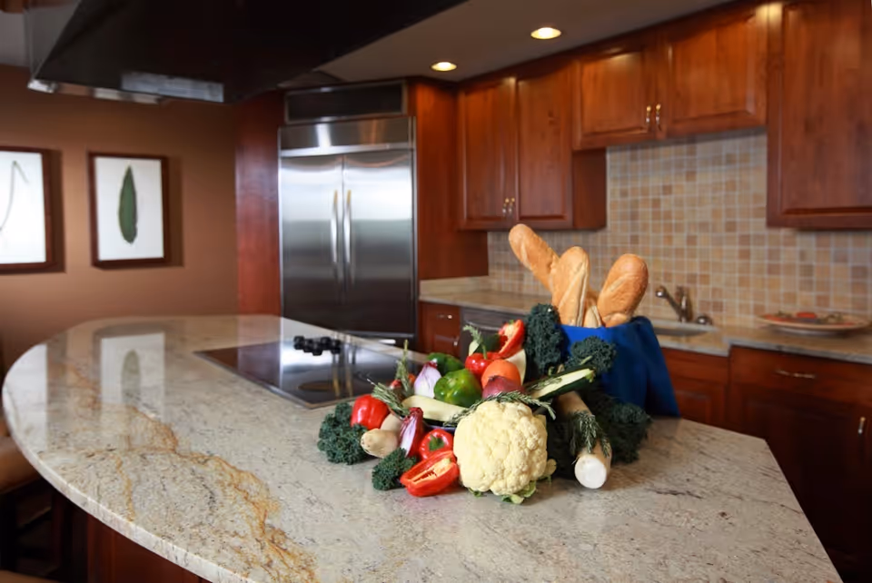 Modern kitchen with a large granite island topped with fresh vegetables and baguettes, wooden cabinets, and a stainless steel refrigerator in the background.