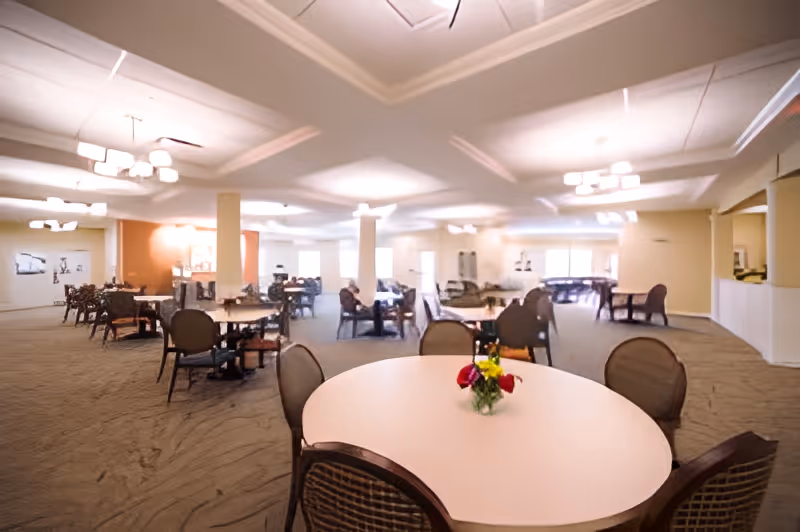 A spacious dining room in a senior living facility with multiple round tables and chairs arranged throughout the room. The tables have small floral centerpieces, and the room is well-lit with ceiling lights and natural light from windows. The carpeted floor and neutral-colored walls create a warm and inviting atmosphere.