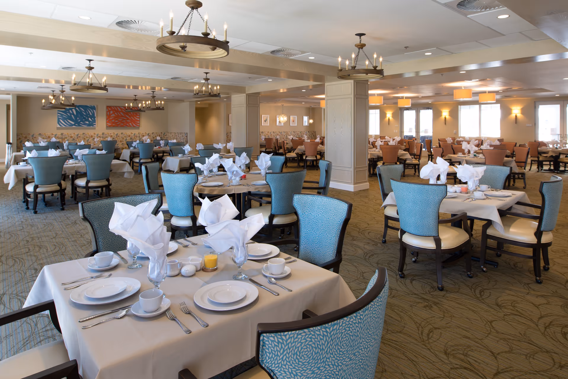 Spacious dining room with multiple tables set with white tablecloths, plates, cups, utensils, and folded white napkins. The room features blue and brown upholstered chairs, carpeted floor with a subtle pattern, and decorative chandeliers hanging from the ceiling. Large windows allow natural light to brighten the space.