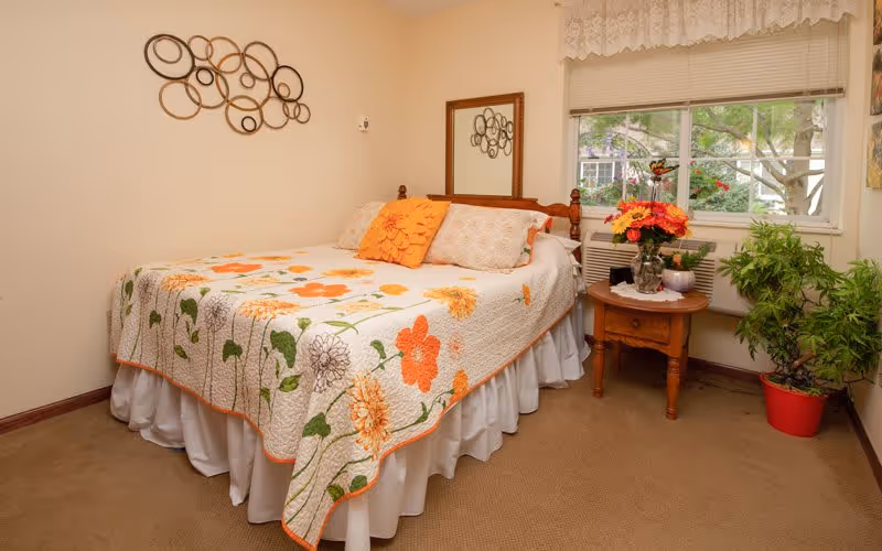 A small bedroom with a bed covered by a floral quilt, a wooden nightstand holding a vase of flowers, and a window showing trees outside.