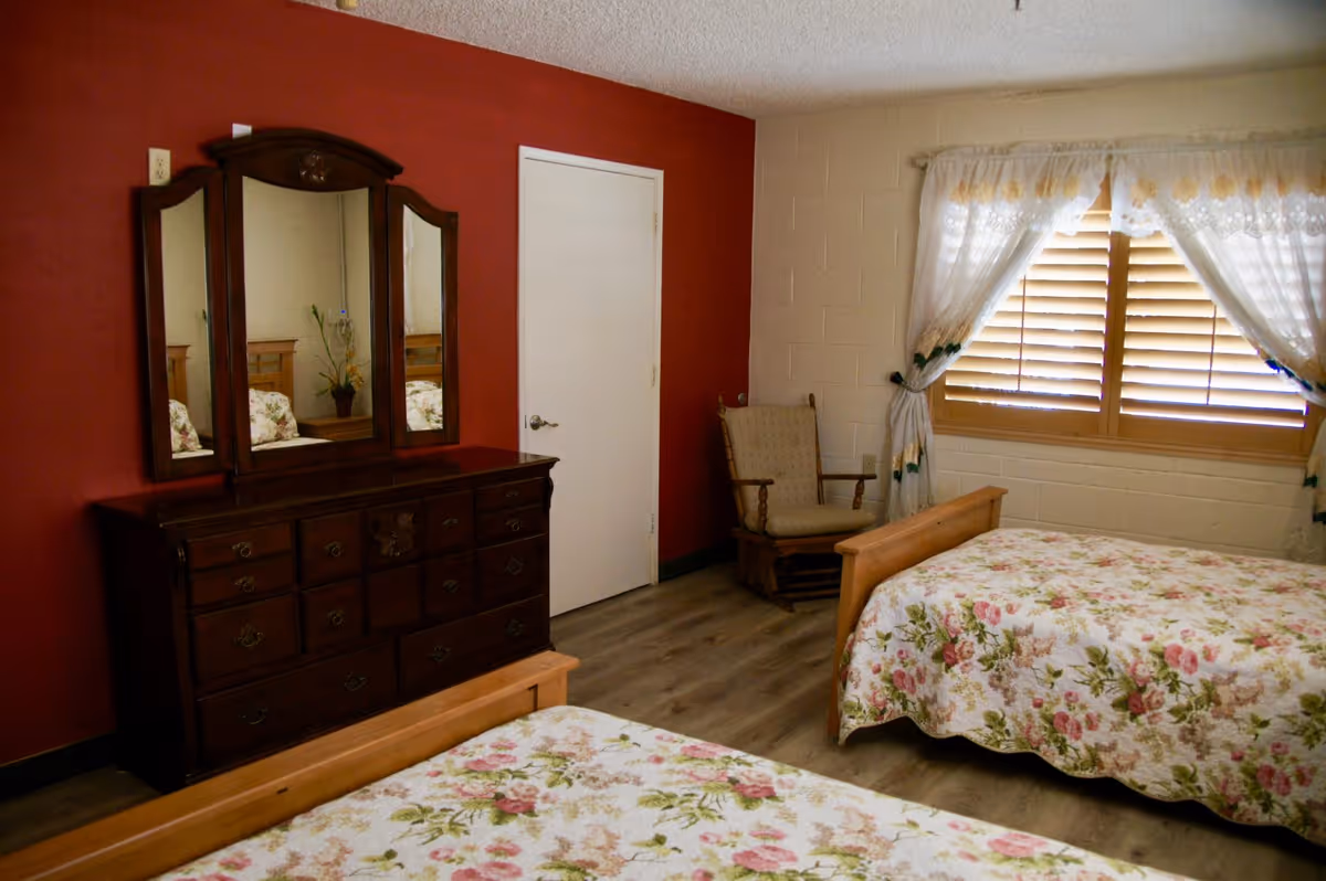 A bedroom with two twin beds covered in floral bedspreads, a wooden dresser with a three-panel mirror against a red accent wall, a white door, a cushioned wooden rocking chair, and a window with wooden blinds and white lace curtains.