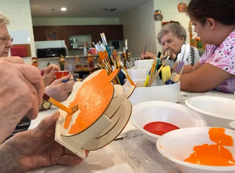 Several elderly individuals and a caregiver are seated around a table engaged in a painting activity. One person is painting an orange pumpkin-shaped wooden craft. The table is filled with paintbrushes, paint containers with red and orange paint, and other art supplies. The background shows a kitchen area with cabinets and appliances.