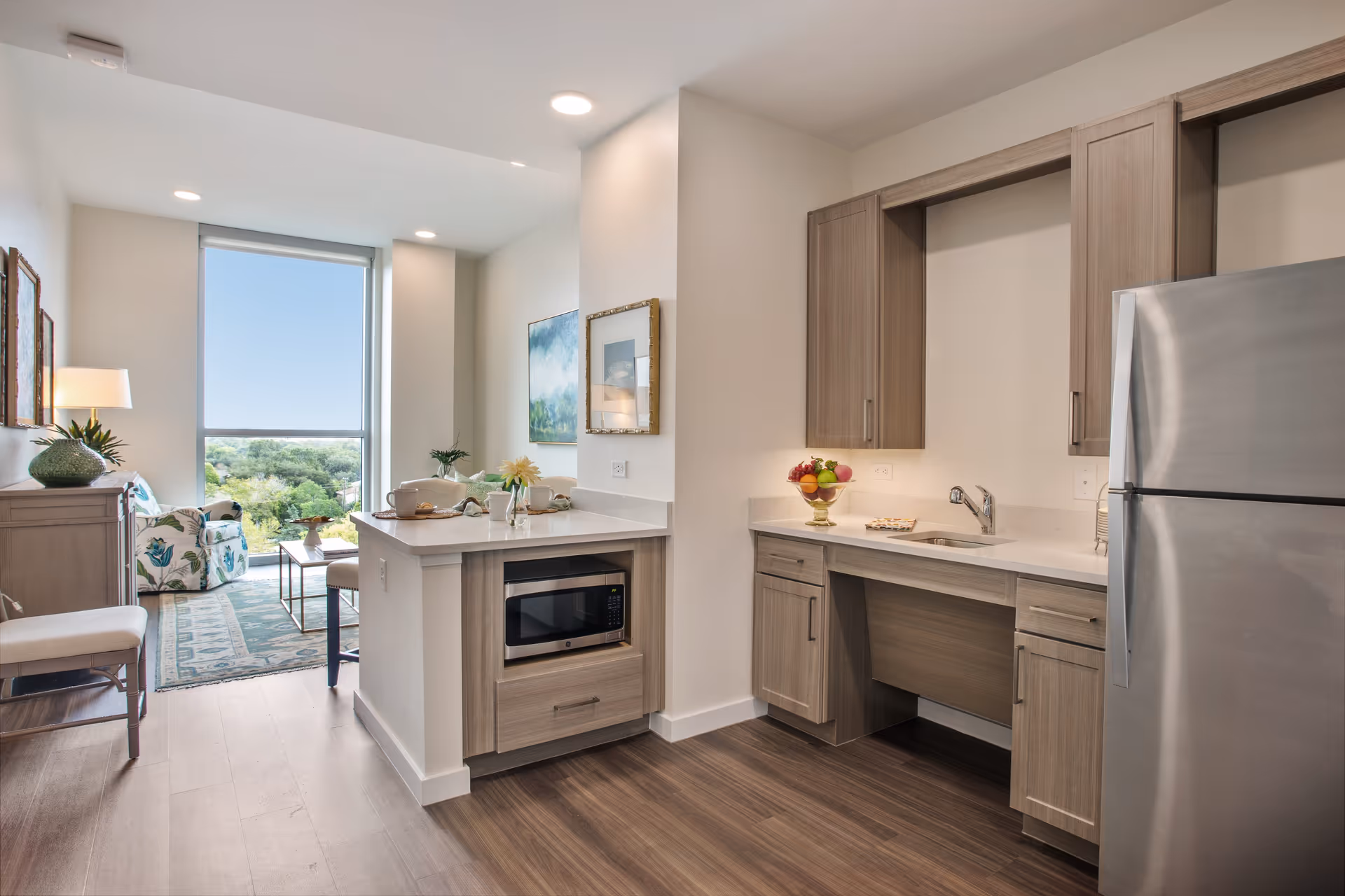 A modern kitchen area with light wood cabinets, a stainless steel refrigerator, a microwave built into the island, and a small sink. The kitchen opens into a living area with a large window showing a view of greenery outside. The living area has a patterned rug, a floral armchair, a coffee table, and a lamp on a side table.