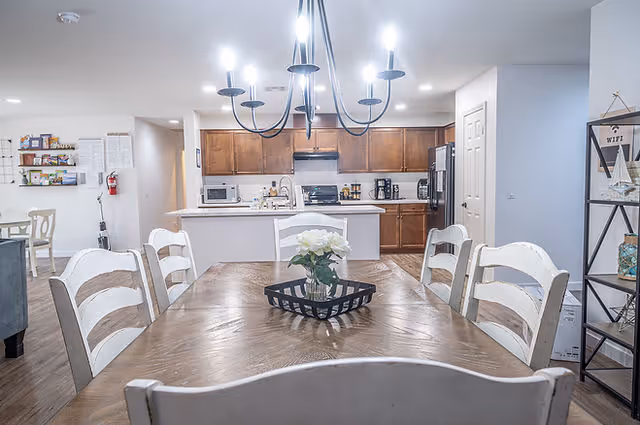 A bright and clean dining area with a wooden table and six white chairs. A black chandelier with six lights hangs above the table. In the background, there is a kitchen with wooden cabinets, a microwave, stove, coffee maker, and refrigerator. To the right, a black metal shelf holds decorative items and a small sign that says 'WIFI'.
