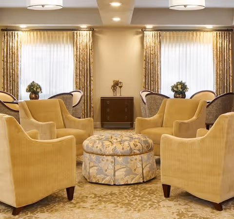 A cozy assisted-living lounge with four yellow armchairs arranged around a patterned round ottoman, with curtained windows and a small cabinet in the background.