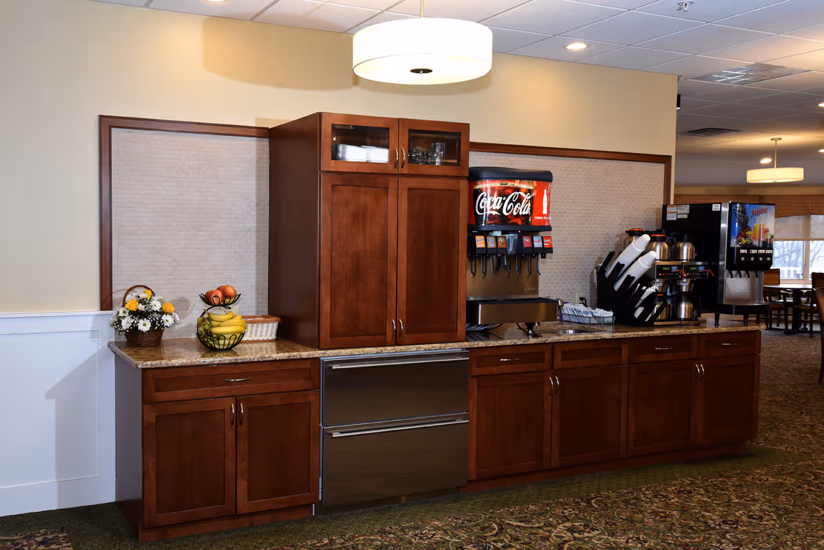 A beverage station in a senior living facility with wooden cabinets, a Coca-Cola soda dispenser, a juice dispenser, coffee pots, cups, and a basket of fruit and flowers on the countertop. The area is well-lit with ceiling lights and has a patterned carpet floor.