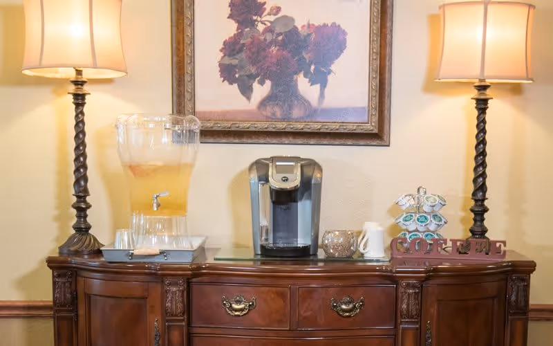 A wooden sideboard with two lit table lamps on each end, a coffee maker in the center, a glass beverage dispenser with cups on the left, and a decorative sign spelling 'COFFEE' on the right. Above the sideboard is a framed painting of flowers.