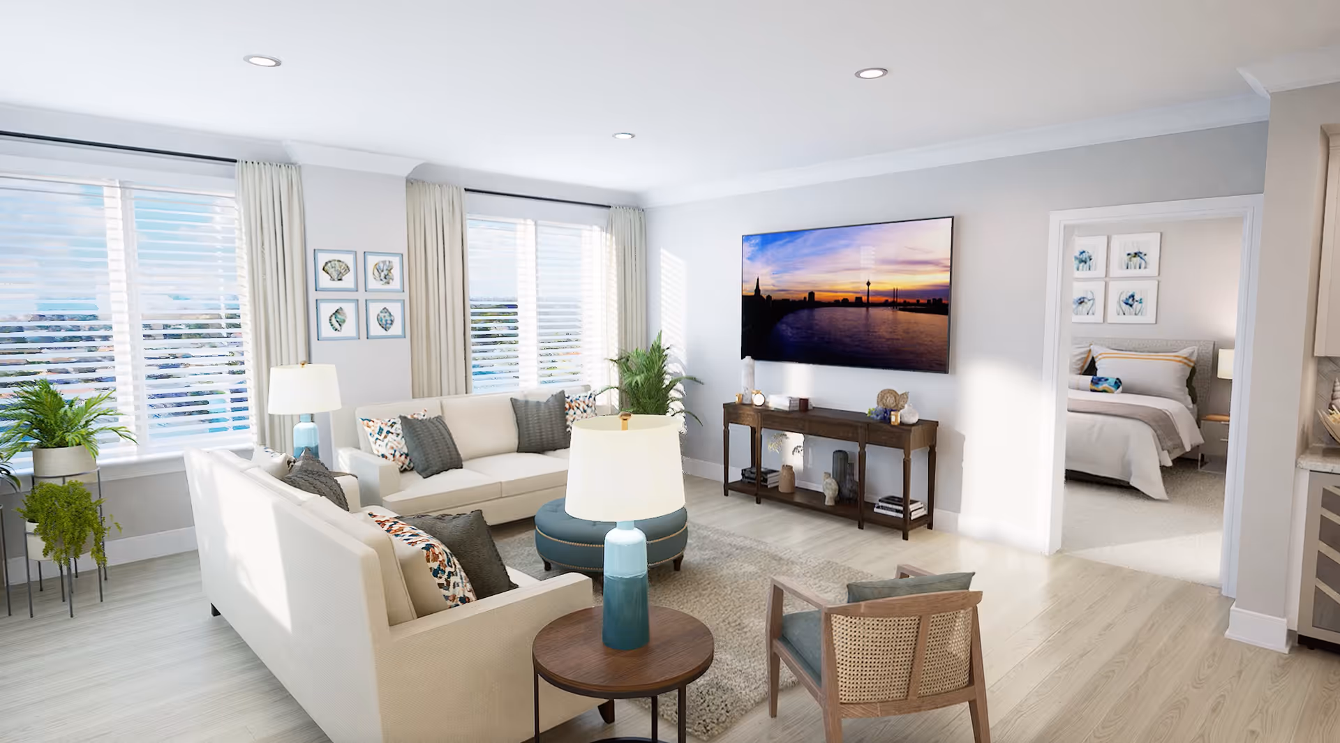 A bright and modern living room with two beige sofas, a wooden armchair, a round ottoman, and a side table with a blue lamp. Large windows with white blinds and cream curtains let in natural light. A flat-screen TV is mounted on the wall above a wooden console table decorated with small plants and ornaments. Through an open doorway, a bedroom with a neatly made bed and framed artwork on the wall is visible.