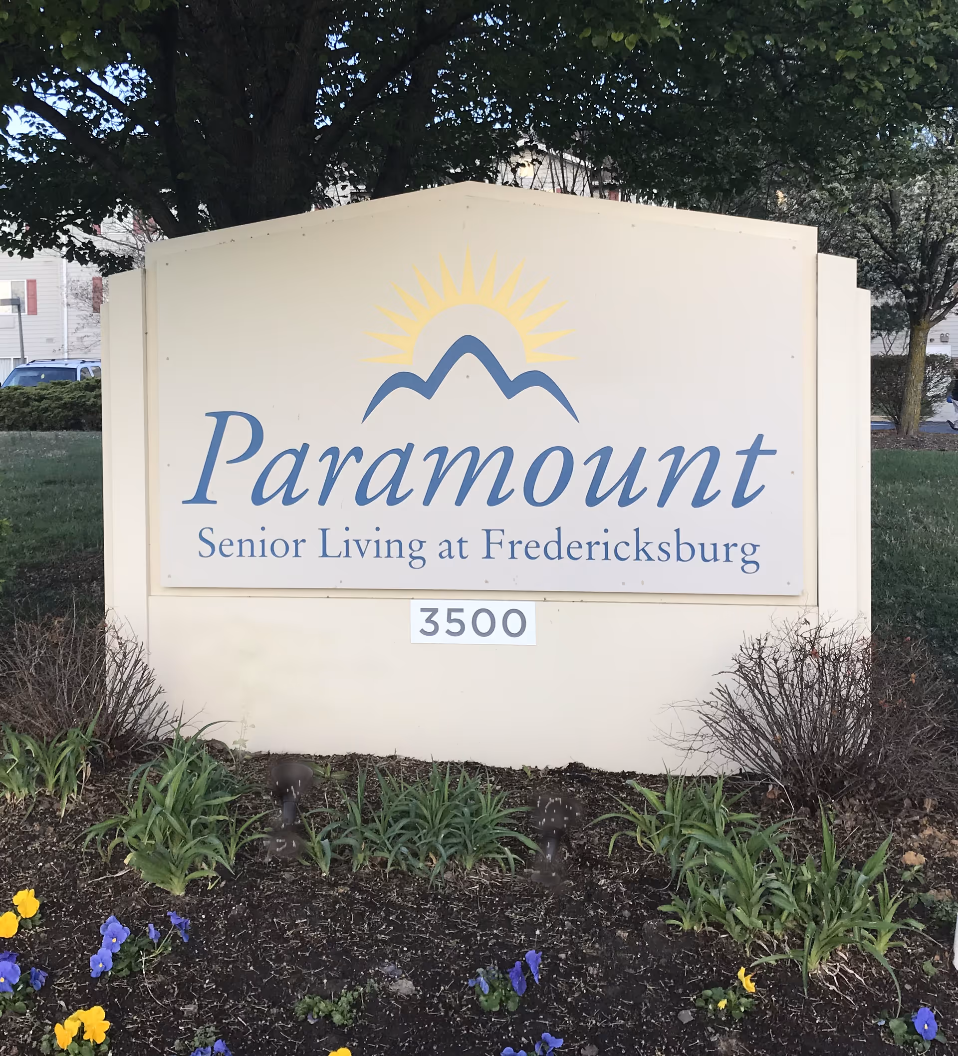 Outdoor sign for Paramount Senior Living at Fredericksburg with the address number 3500, surrounded by plants and flowers, with trees and a building in the background.