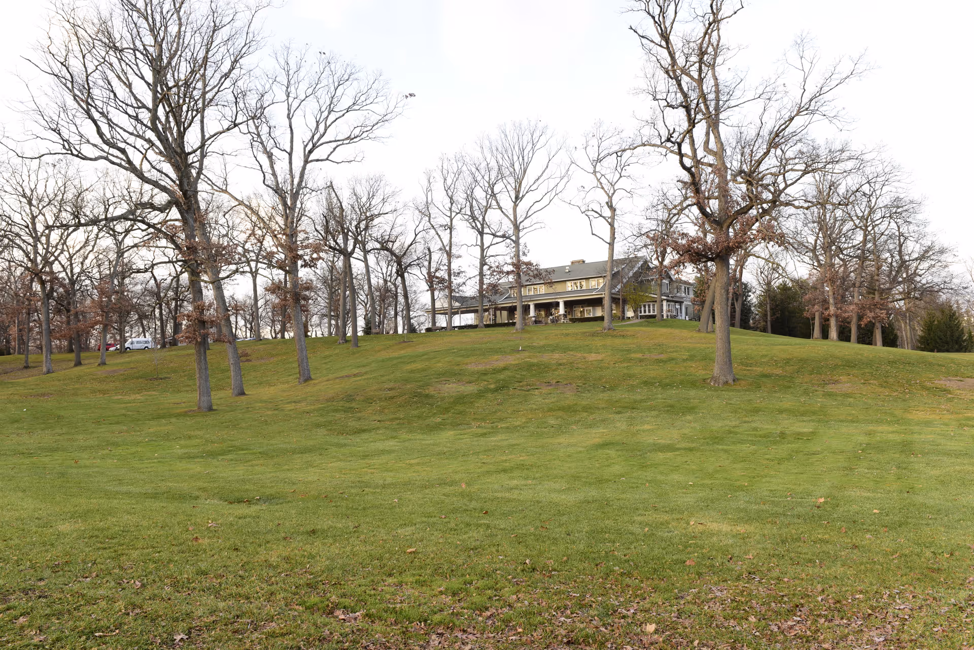 A large grassy hill with scattered leafless trees leading up to a two-story building with a porch, set against a pale sky.