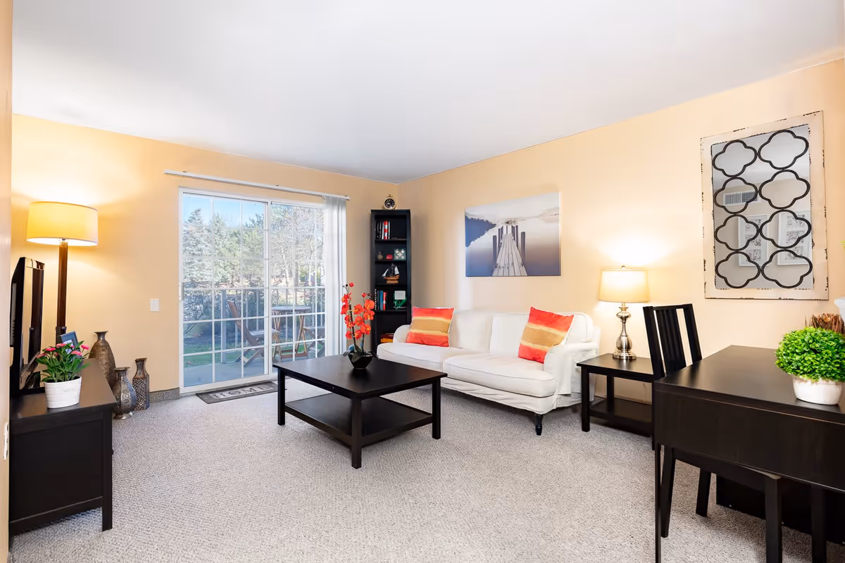 A bright living room with beige walls and carpeted floor featuring a white sofa with colorful cushions, a dark wooden coffee table, a matching side table with a lamp, a black bookshelf, a TV on a stand, a floor lamp, and a sliding glass door leading to an outdoor area.
