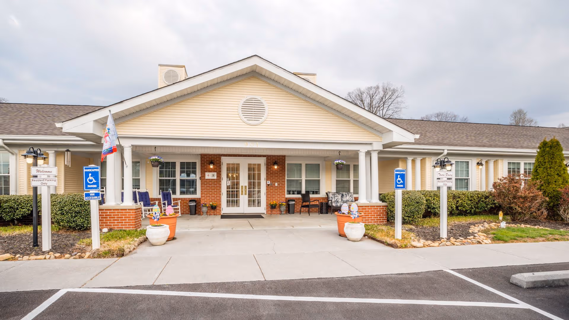 Front exterior view of American House Lebanon facility showing a single-story building with a covered entrance supported by white columns. There are two handicap parking signs and a welcome sign near the entrance. The area is landscaped with bushes and potted plants, and there are chairs on the porch.