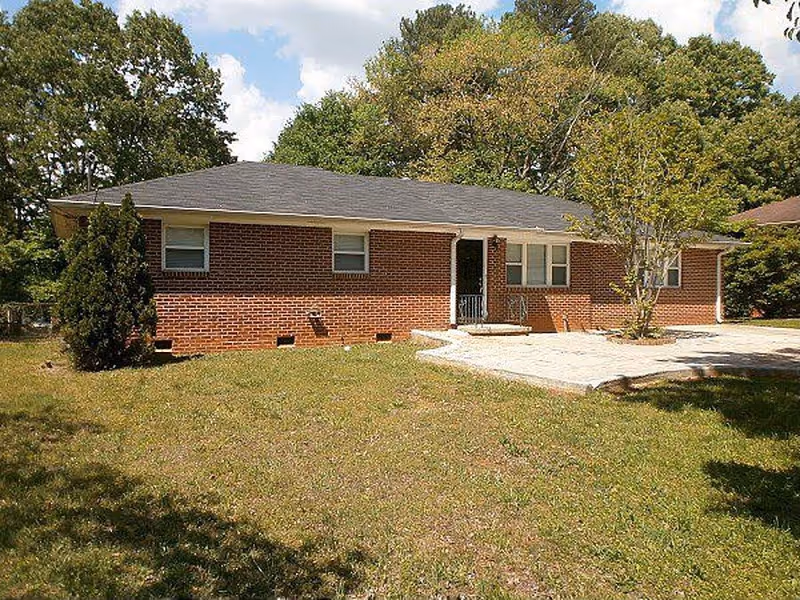 Single-story brick building with a dark shingled roof, several windows, a small front porch with steps, and a paved area in front. The building is surrounded by grass and trees under a partly cloudy sky.