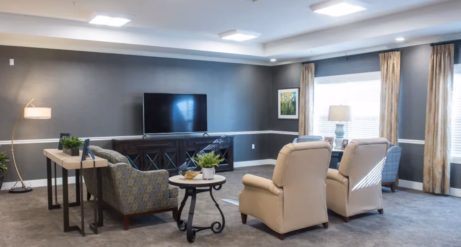 A well-lit living room in a senior living facility with two beige armchairs, a patterned sofa, a round side table with a plant, a wooden console with a flat-screen TV, a floor lamp, and large windows with beige curtains.