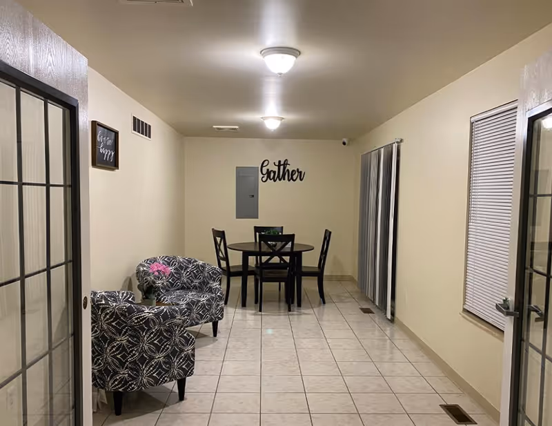 A narrow room with beige walls and tiled floor featuring a small round dining table with four black chairs in the center. On the left side, there are two patterned armchairs with a small table holding a pink flower. The wall at the far end has a decorative sign that says 'Gather'. There are two ceiling lights, a window with blinds on the right, and sliding glass doors on both sides.