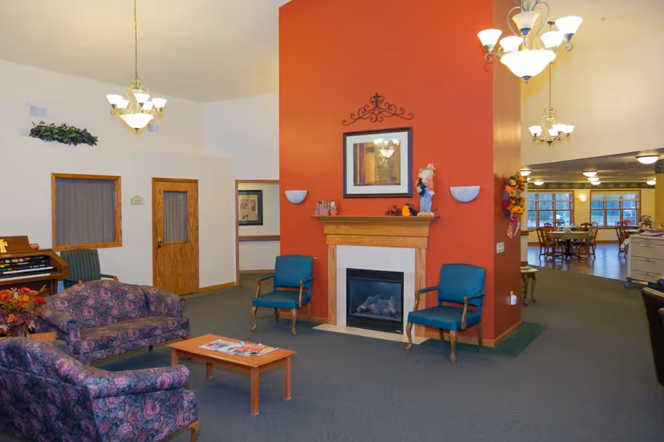 A bright assisted-living common room with sofas and armchairs around a coffee table, a central fireplace with mantel, and a dining area visible in the background.