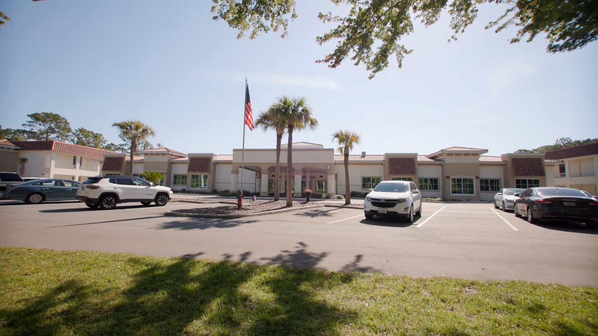 Front exterior view of Indigo Manor Nursing and Rehabilitation - Bedrock Healthcare building with a parking lot in front, several parked cars, palm trees, and an American flag on a flagpole under a clear sky.