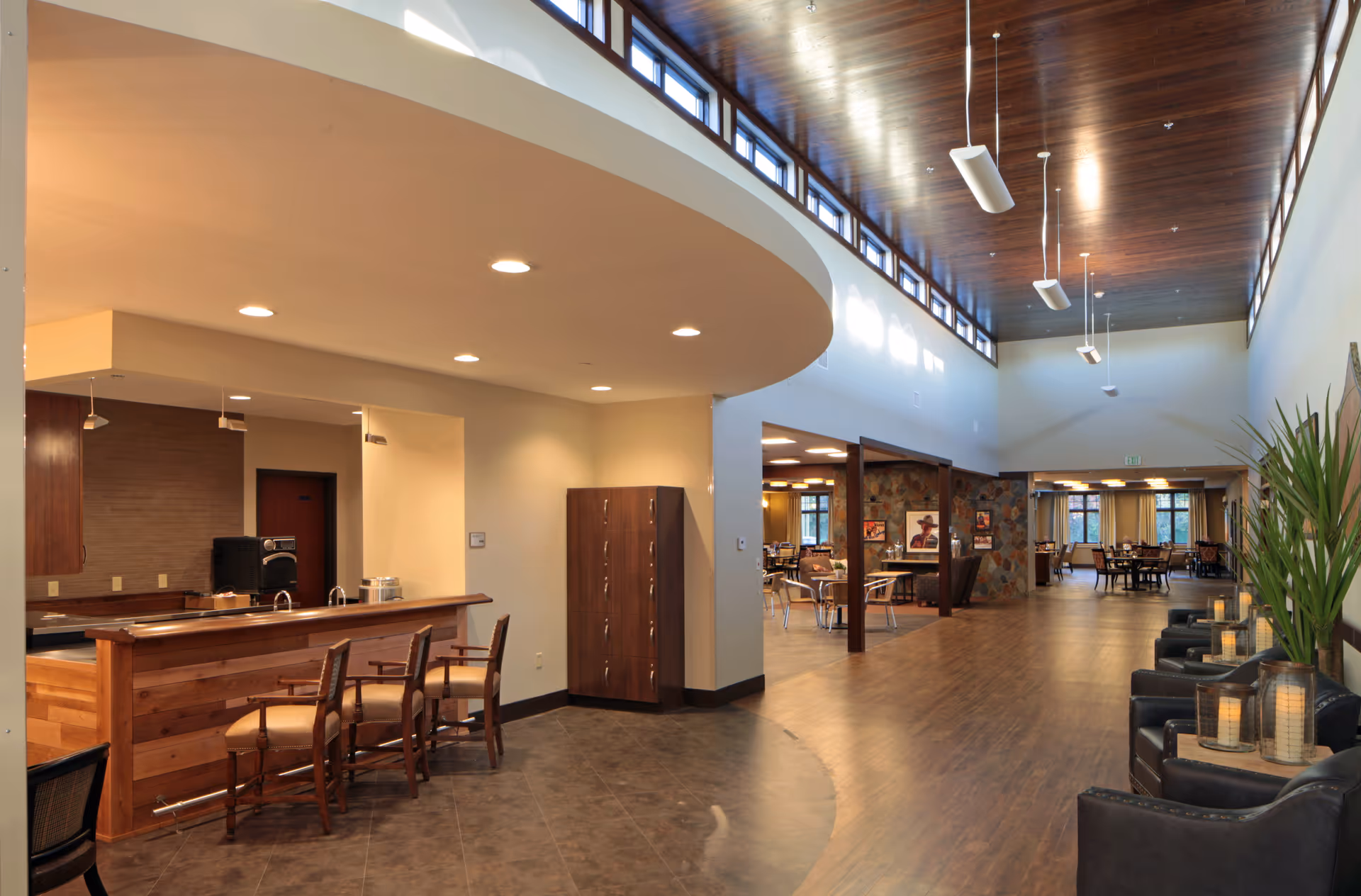 Interior view of a senior living facility showing a spacious common area with a wooden ceiling and high windows. On the left, there is a kitchen counter with three chairs. The area extends into a dining space with tables and chairs, and there are comfortable black armchairs with candles on the right side. The space is well-lit with recessed lighting and pendant lights.