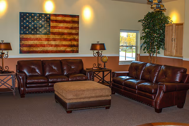 A cozy living room with two dark brown leather sofas facing each other, a large square ottoman in the center, two side tables with lamps, a large American flag artwork on the wall, a window showing outdoor greenery, and a tall potted plant in the corner.