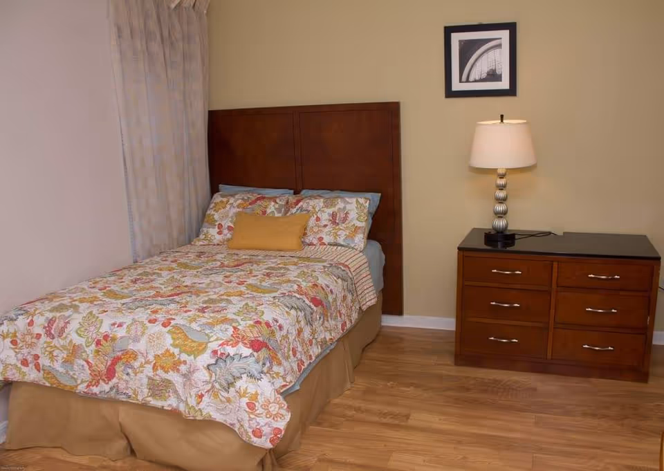 A bedroom with a bed featuring a floral patterned bedspread and pillows, a wooden headboard, a wooden dresser with six drawers, and a table lamp with a white shade. A framed black and white photo hangs on the beige wall above the dresser. Light-colored curtains cover the window on the left side.