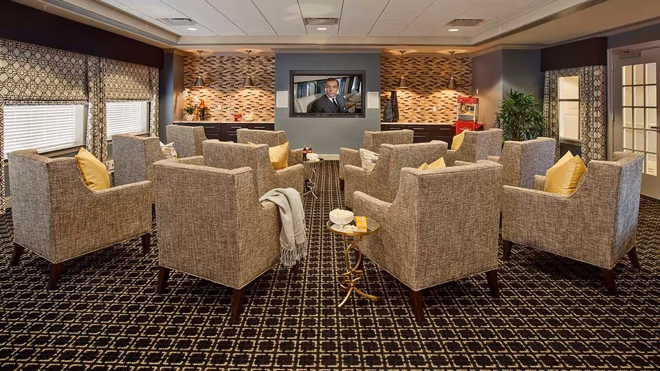 A cozy lounge area with multiple gray upholstered armchairs arranged in rows facing a wall-mounted flat screen TV. The room features patterned carpet, decorative window treatments, and a small side table with popcorn and drinks. The back wall has a mosaic tile backsplash with two wall lamps and a popcorn machine on the right side.