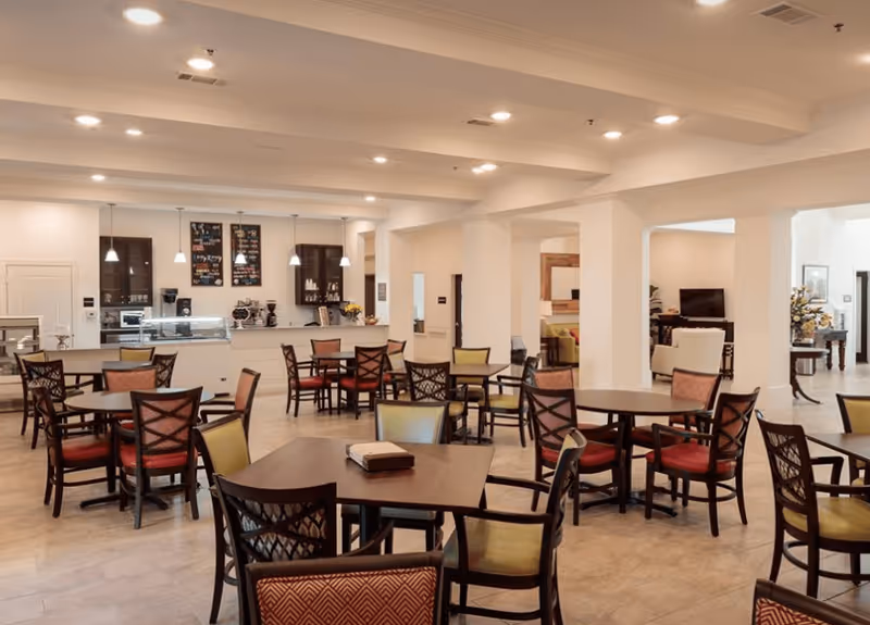 A spacious dining room with multiple dark wood tables and chairs with red and green cushions. The room has a light-colored tile floor and white walls with ceiling lights. In the background, there is a counter area with hanging pendant lights, coffee machines, and menu boards on the wall. Adjacent to the dining area, there is a living room space with sofas and a television.