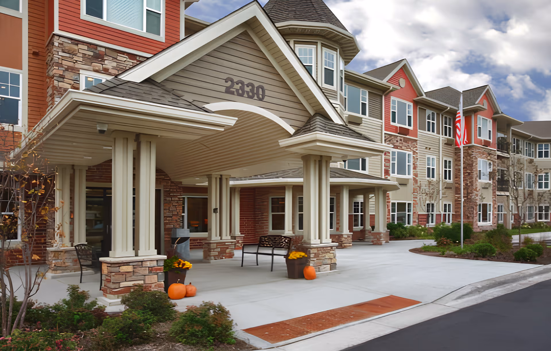 Exterior view of Bel Rae Senior Living facility showing the main entrance with a covered driveway supported by columns. The building has multiple floors with a mix of stone and siding in earth tones and red accents. There are pumpkins and potted flowers near the entrance, and an American flag is visible on a flagpole to the right.