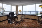 A cozy interior room with large windows offering a panoramic view of a body of water and distant hills. The room features a patterned carpet, wooden chairs with upholstered seats, a table with a computer monitor, a floor lamp, and a hanging light fixture.